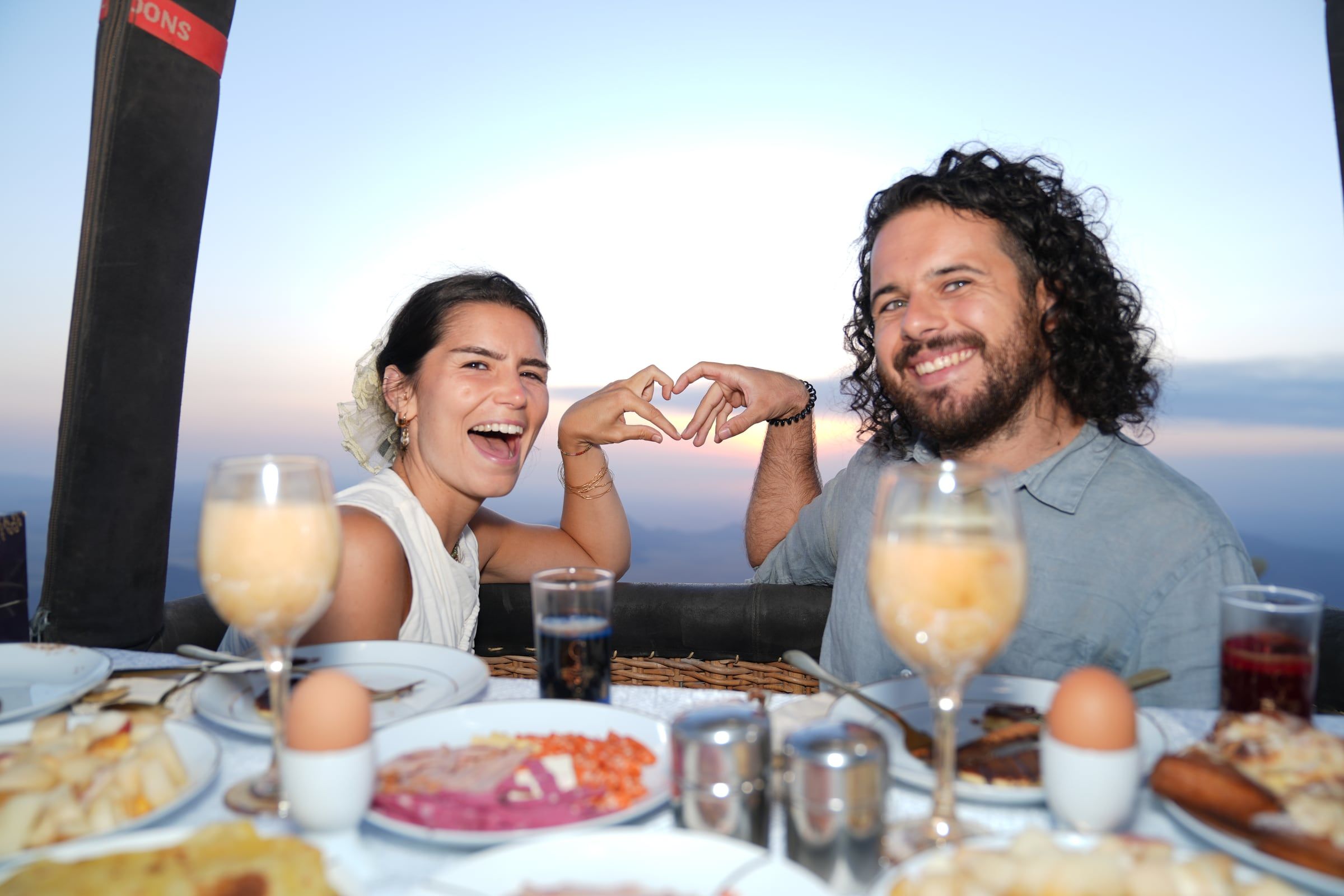 Laughing couple making a heart with their hands at a sunrise breakfast in the balloon