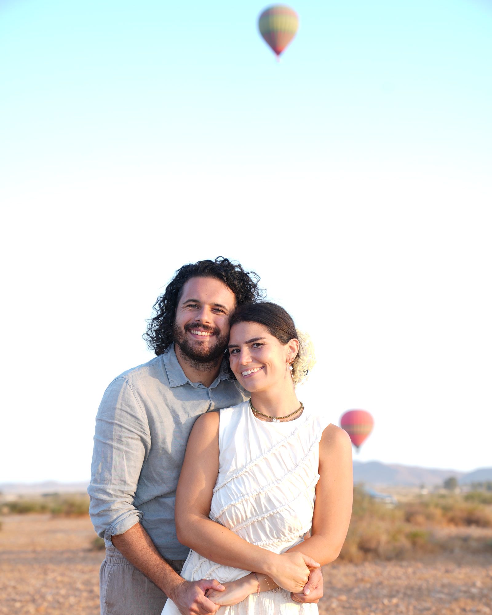 Couple embracing during an engagement photoshoot in the Agafay desert near Marrakech, hot-air balloons in the sky